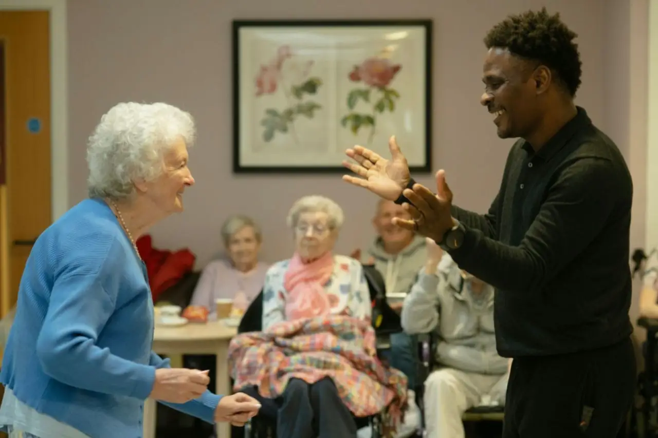 Jeune homme qui danse en frappant dans ses mains avec une dame âgée souriante, dans la salle commune d'une maison de retraite