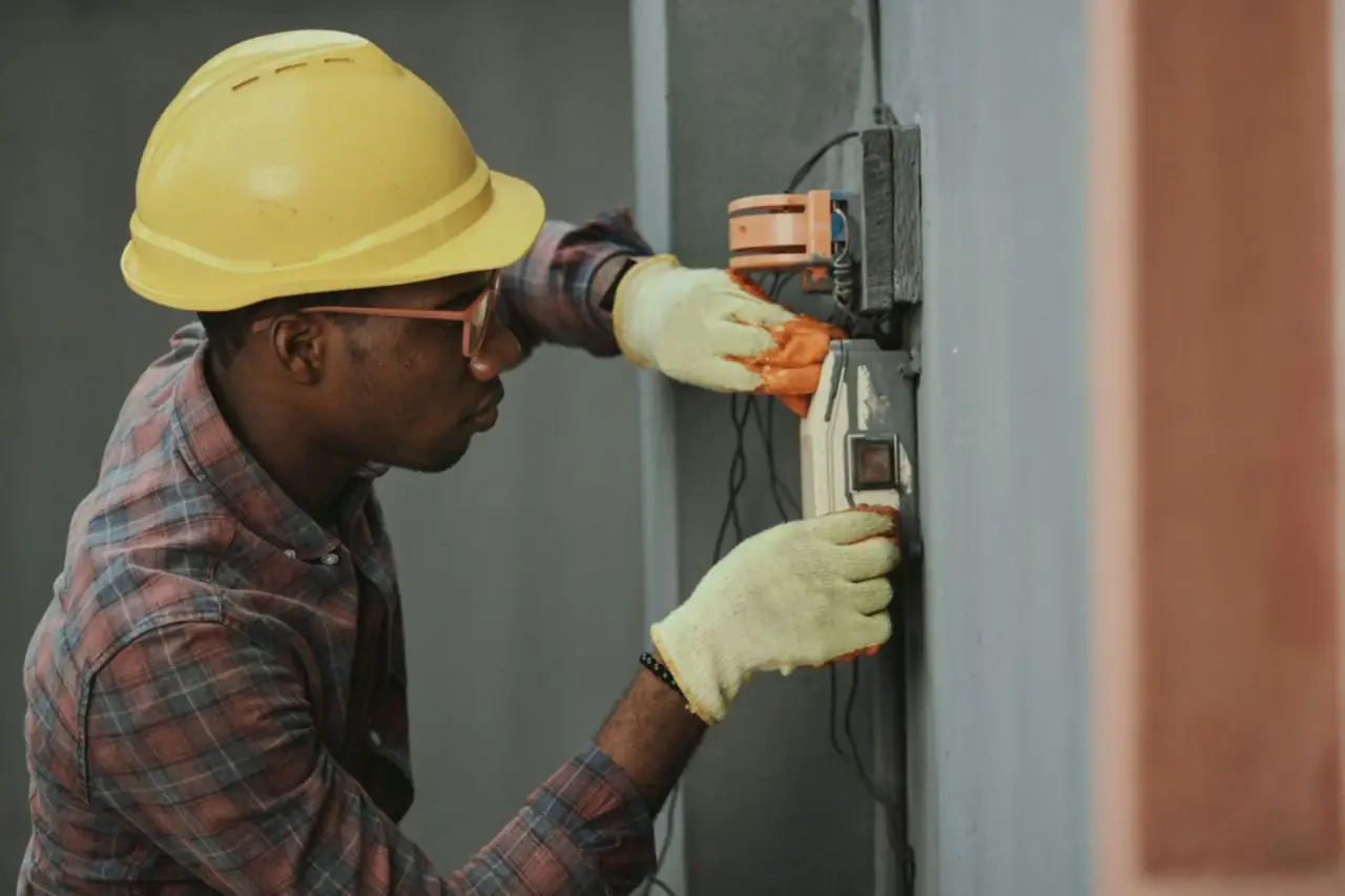Jeune électricien en casque de chantier jaune et gants de travail qui installe un boîtier électrique au mur