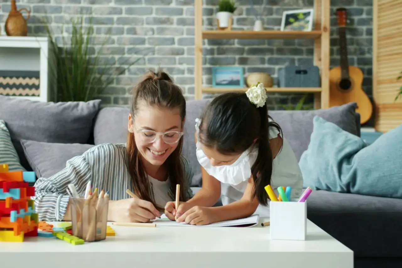 Jeune femme souriante qui dessine avec une petite fille sur la table basse d'un salon, entourées de crayons et de jeux de construction