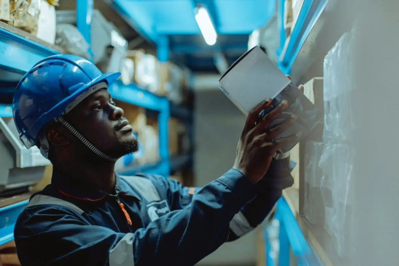 Magasinier en casque de sécurité bleu et tenue de travail qui range un carton sur une étagère d'entrepôt