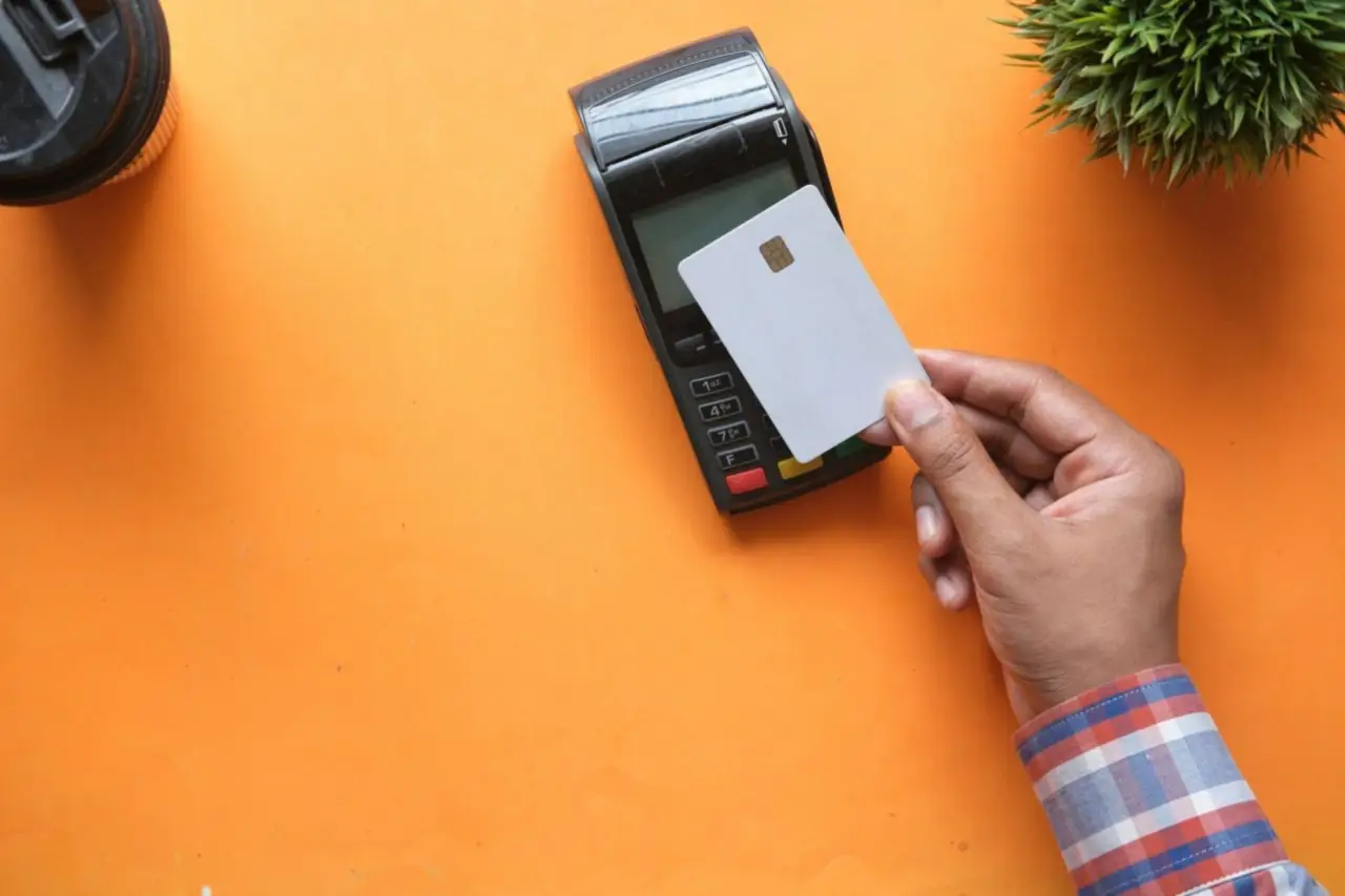 Main qui approche une carte bancaire vierge d'un terminal de paiement posé sur une table orange