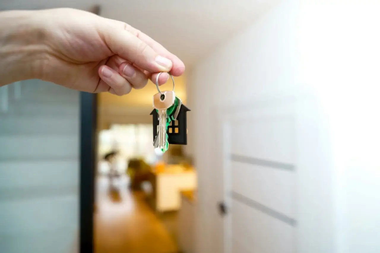 Main qui tend un trousseau de clés avec un porte-clés en forme de maison, devant l'entrée d'un appartement lumineux